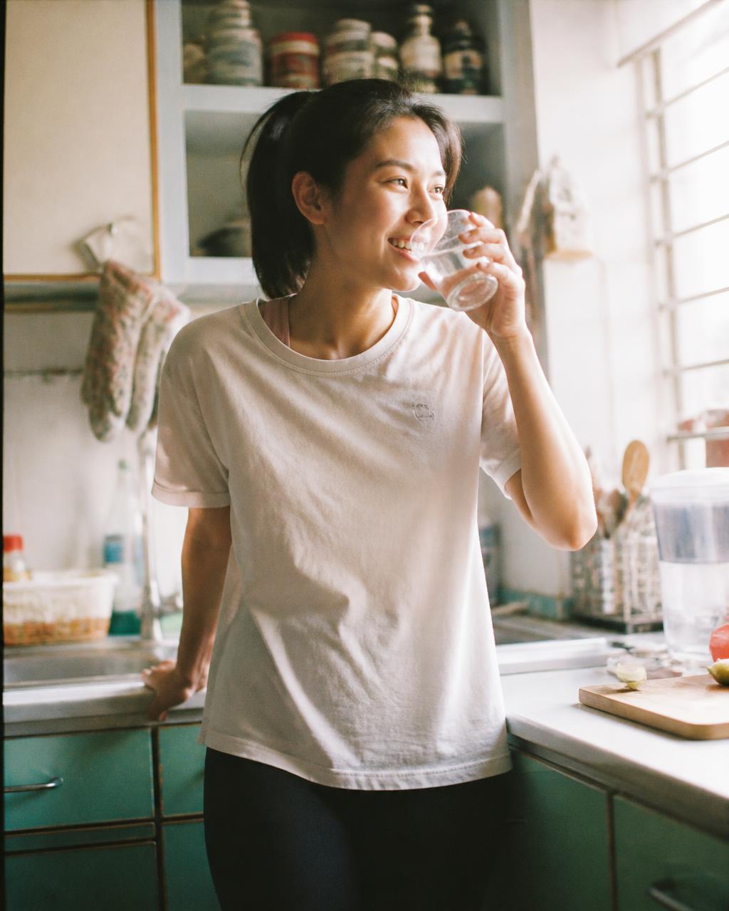 A young Singaporean woman drinking water in her sunlit HDB kitchen after a morning run